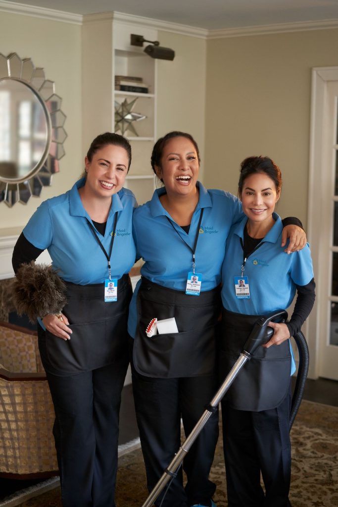 Three professional Maid Brigade team members in blue uniforms smiling together after cleaning a home, representing teamwork and trust in professional housecleaning.