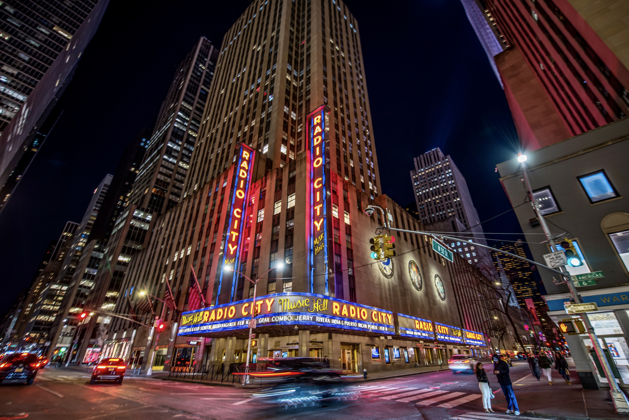 Radio City Music Hall lit up at night in New York City, with traffic and pedestrians on the street.